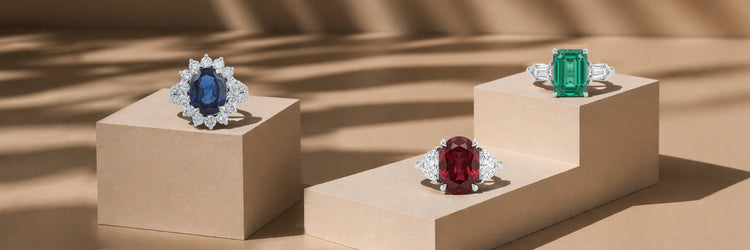 Three gemstone engagement rings in white gold on beige pedestals — oval blue sapphire with diamond halo, cushion cut ruby with trillion diamond three-stone setting, and emerald cut green emerald with baguette side stones