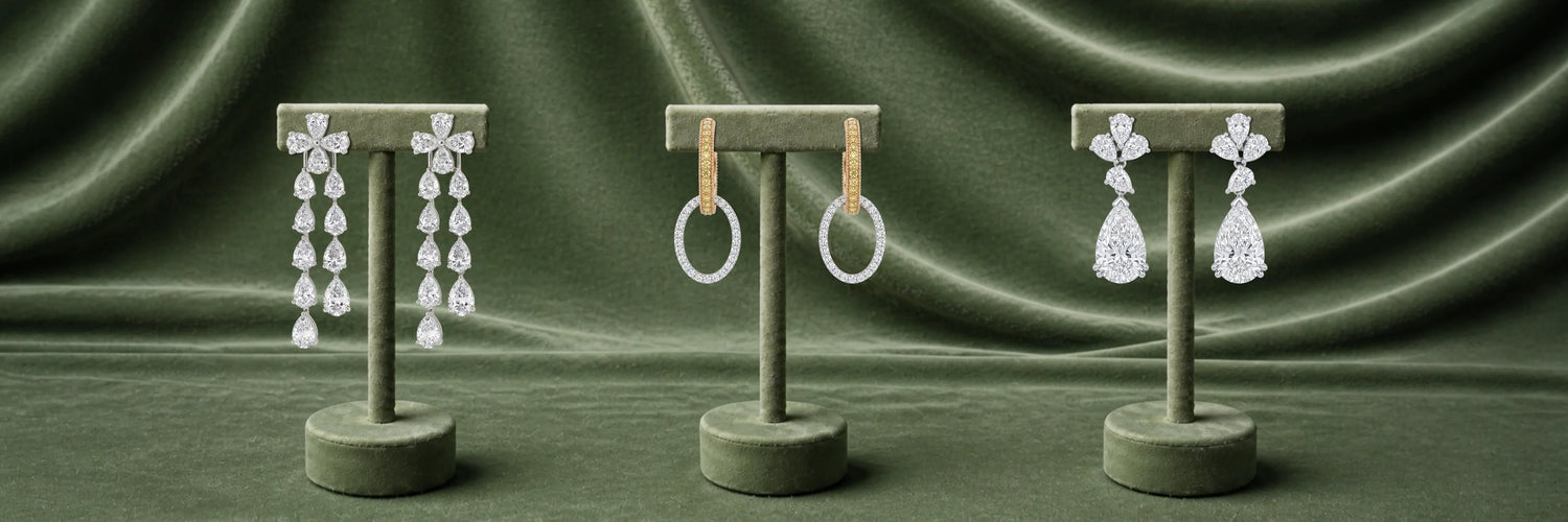 Three pairs of lab-grown diamond earrings on olive stands — white gold pear cut cascading chandelier drop earrings, yellow gold pavé oval hoop earrings, and white gold triple pear cluster with large pear drop earrings on green satin
