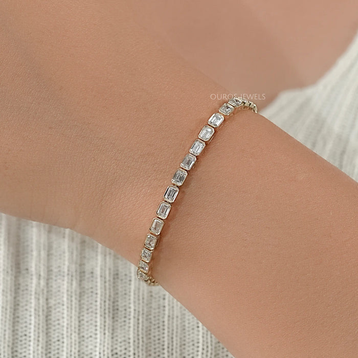east west set emrald cut man made diamonds bezel set tennis bracelet in yellow gold on hand